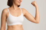A woman in a white sports bra flexes her right arm, showing her bicep and highlighting the potential arm lift surgery benefits against a neutral background.