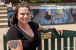 A woman with tattoos sits on a green bench outdoors, smiling at the camera. Behind her is a mural with graffiti art, including the word "peace" and a peace sign—she radiates confidence, like someone who knows how to choose skin care products.
