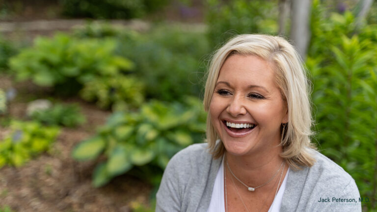 A woman with blonde hair and a gray cardigan smiles while sitting outdoors in a garden setting, showing off her confidence after trying the best cosmetic injectables for lips.