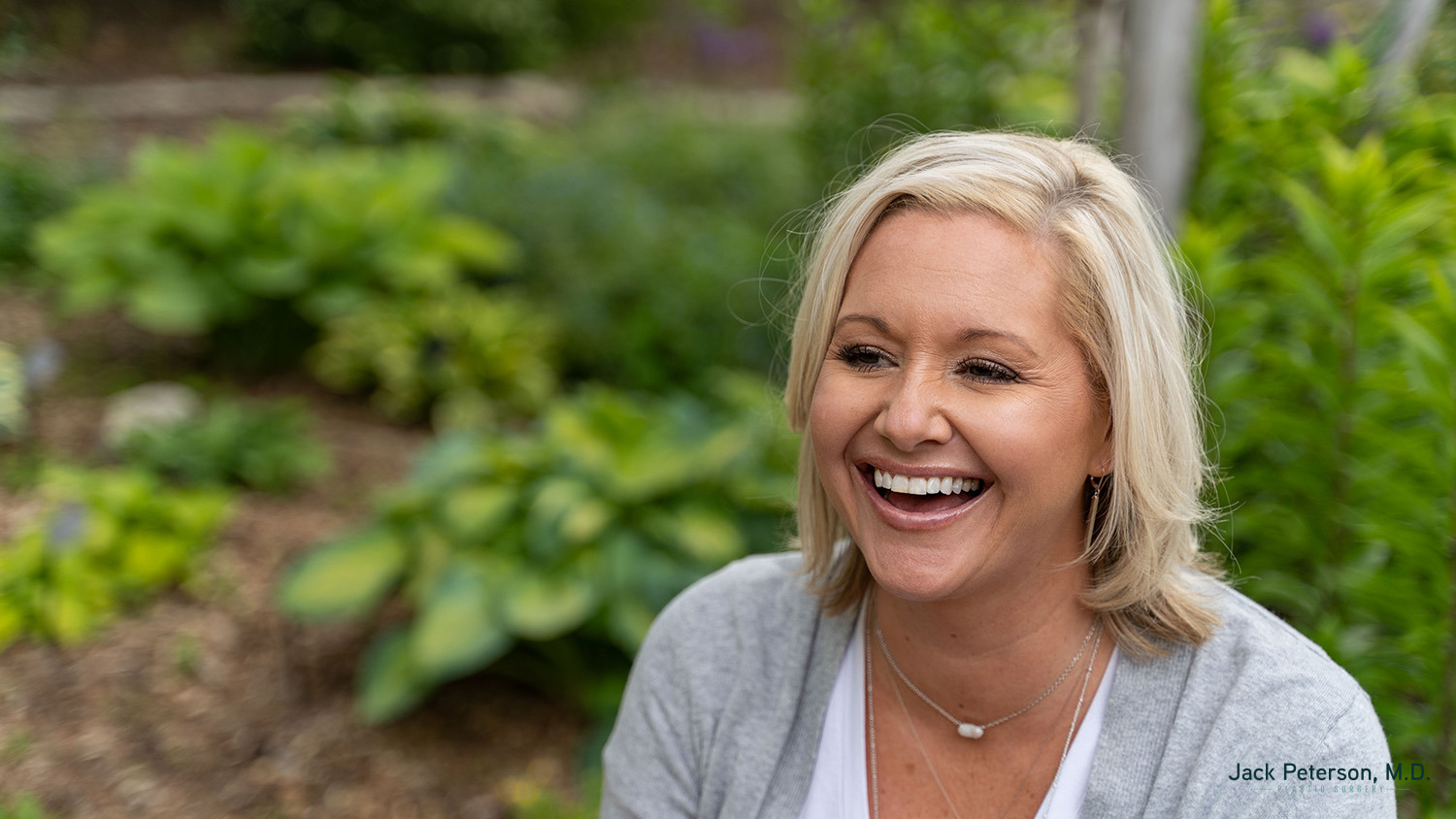 A woman with blonde hair and a gray cardigan smiles while sitting outdoors in a garden setting, showing off her confidence after trying the best cosmetic injectables for lips.