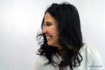 Woman with long dark hair smiling and looking to her left, wearing a light textured top against a plain white background, showcasing confidence that rivals the results of the best cosmetic injectables for lips.