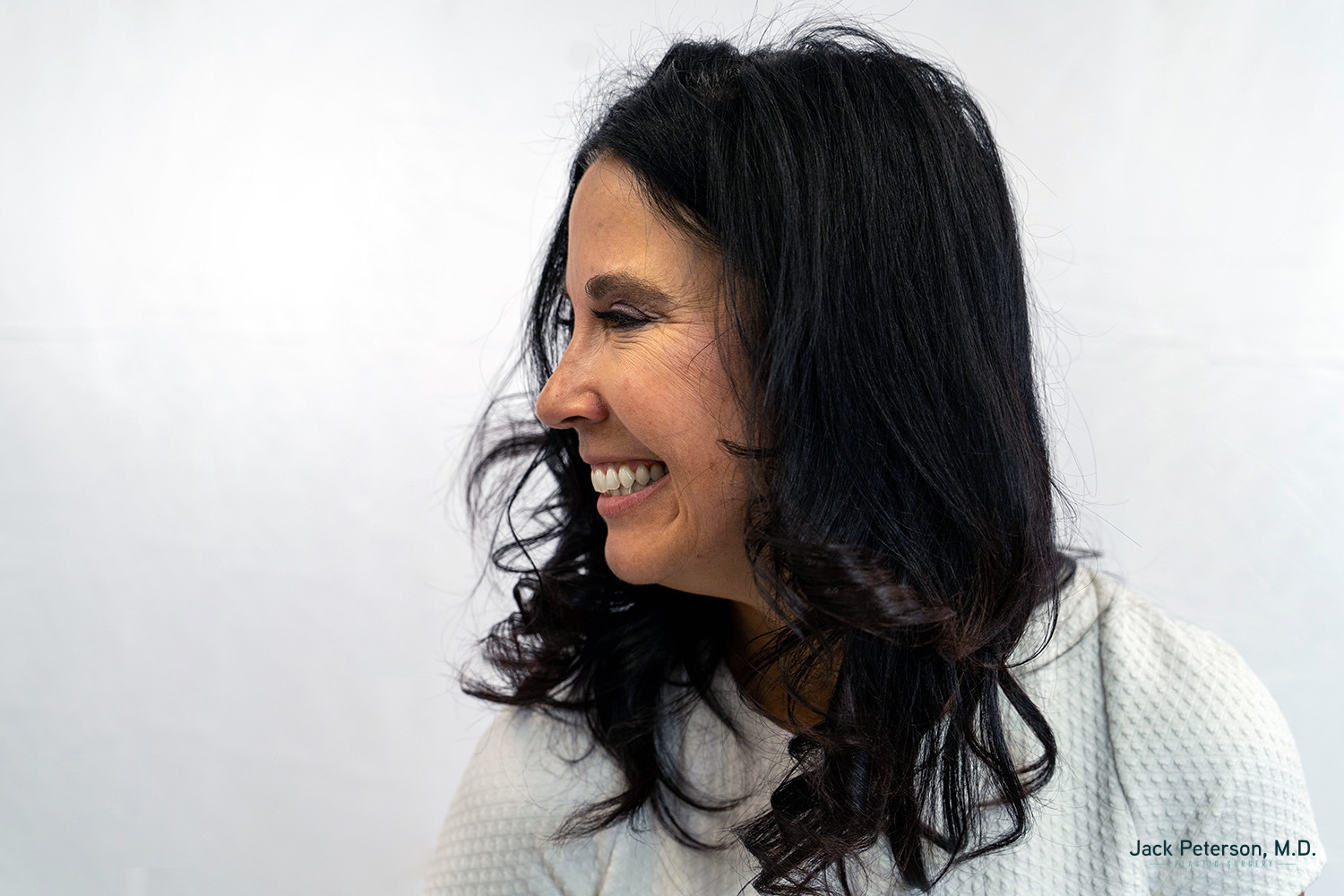 Woman with long dark hair smiling and looking to her left, wearing a light textured top against a plain white background, showcasing confidence that rivals the results of the best cosmetic injectables for lips.